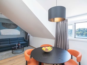 A modern living room with a round table, orange chairs, a dark sofa, a black coffee table, and a large window with a view of trees and water. - Apartments Villa Royale - One Bedroom Apartment With Sea View (Konavle)