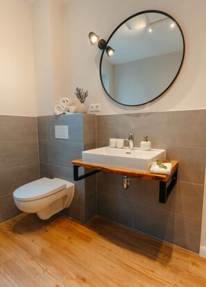 A modern bathroom with a white toilet, a round mirror, and a wooden shelf above the sink. - Fine Stuuv Comfortable Holiday Residence (Ratzeburg)
