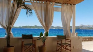 Un espace extérieur aménagé, abrité et doté de deux chaises et d’une table, offrant une vue sur une mer d’un bleu profond et sur une côte lointaine.