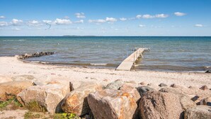 Une plage avec une jetée en béton qui s’avance dans l’eau, entourée de gros rochers et surplombée d’un ciel bleu clair.