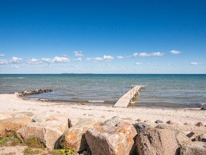 Ein Strand mit einer Betonsteg, der sich ins Wasser erstreckt, umgeben von großen Felsen und einem klaren, blauen Himmel.