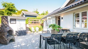 Une terrasse avec un sol en pierre, des sièges d’extérieur, une cheminée en pierre et une maison avec un toit et des fenêtres recouverts de tuiles.