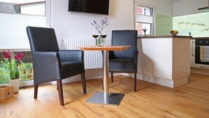 Une cuisine moderne avec une table à manger en bois, deux chaises noires, un téléviseur à écran plat fixé au mur et un petit îlot central doté d’un évier et d’un plan de travail.