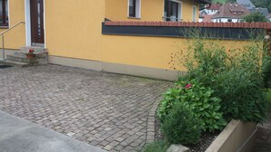 Un bâtiment jaune avec un toit rouge, une allée pavée et un petit jardin agrémenté de plantes et d’un pot de fleurs.