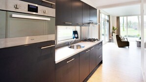 A modern kitchen with dark cabinetry, stainless steel appliances, and a large window providing natural light. - Paviljoen 4 Personen (Zorgvlied)