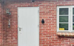 Un edificio de ladrillo rojo con una puerta blanca, una ventana y un letrero amarillo, y una cámara de seguridad negra.