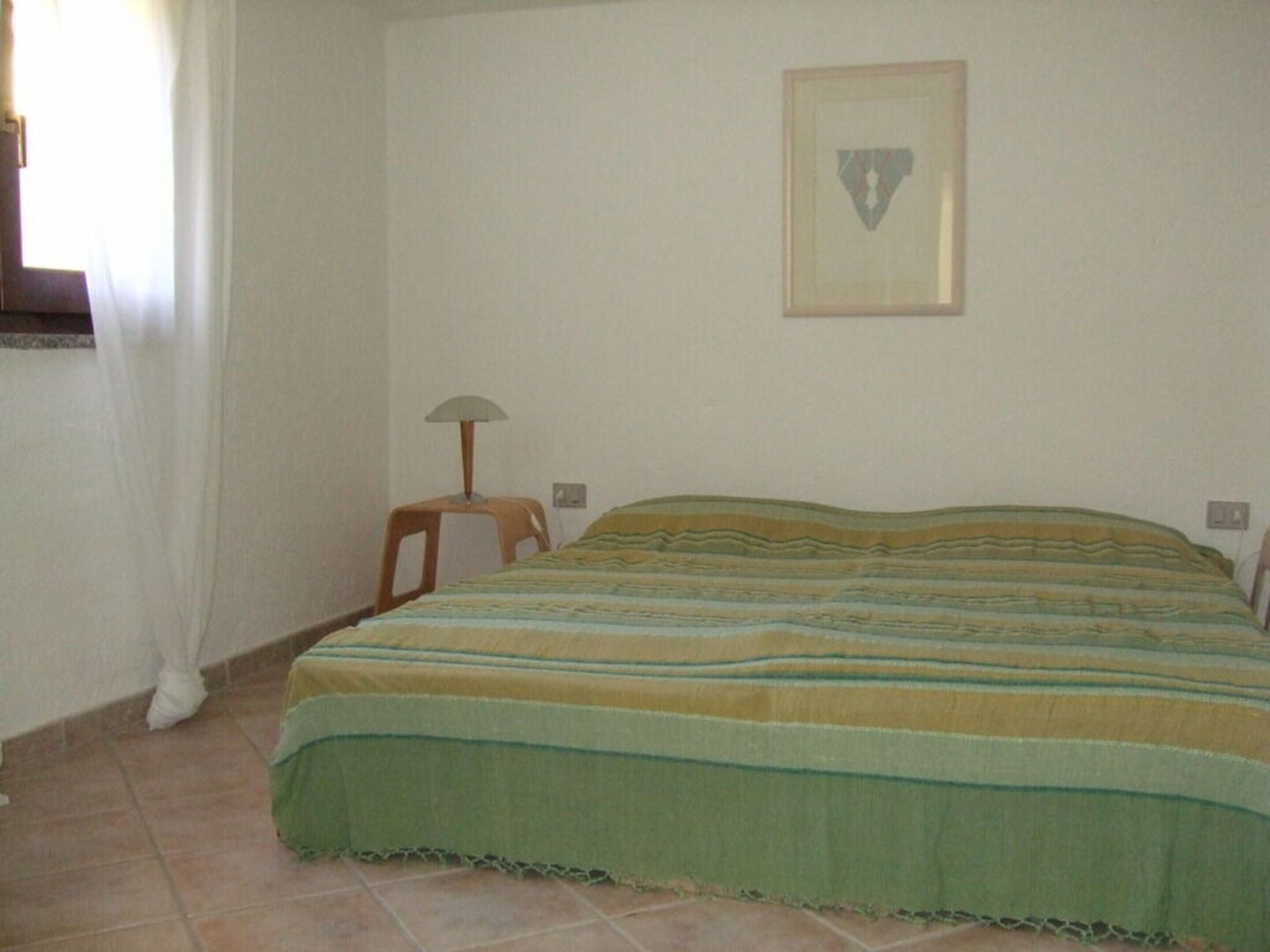 A simple room with a bed, a chair, a lamp, and a framed picture on the wall.