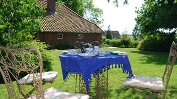 An outdoor dining area with a table set for tea, surrounded by ornate chairs, in a garden with a brick building in the background.