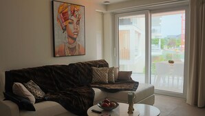A living room with a dark brown sectional sofa, a white armchair, a round coffee table with a bowl of fruit and a candle, and a painting on the wall. - Apartment in Nieuwpoort With sea View (Nieuwpoort)
