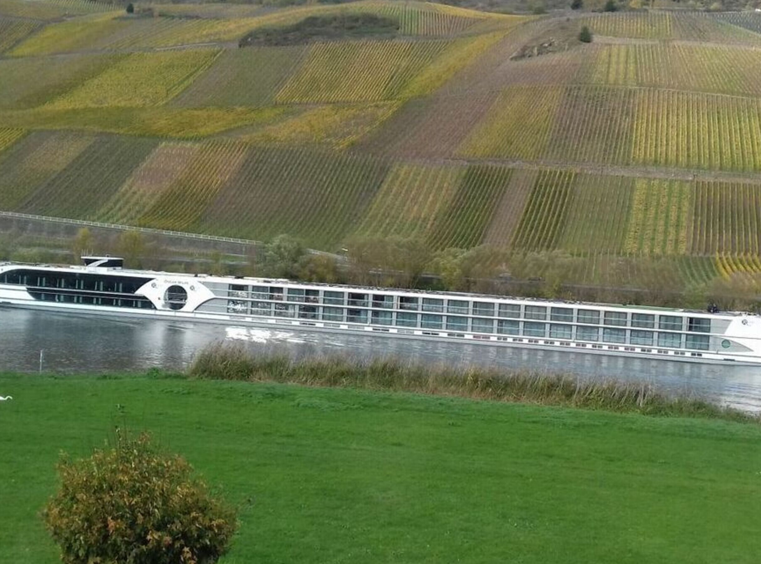 Un barco de crucero fluvial con diseño moderno navegando por un río, con campos verdes y colinas al fondo.