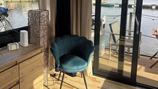 Een moderne kamer met een houten vloer, een blauwgroene stoel, een bureau met een lamp, en uitzicht op een waterpartij met boten.