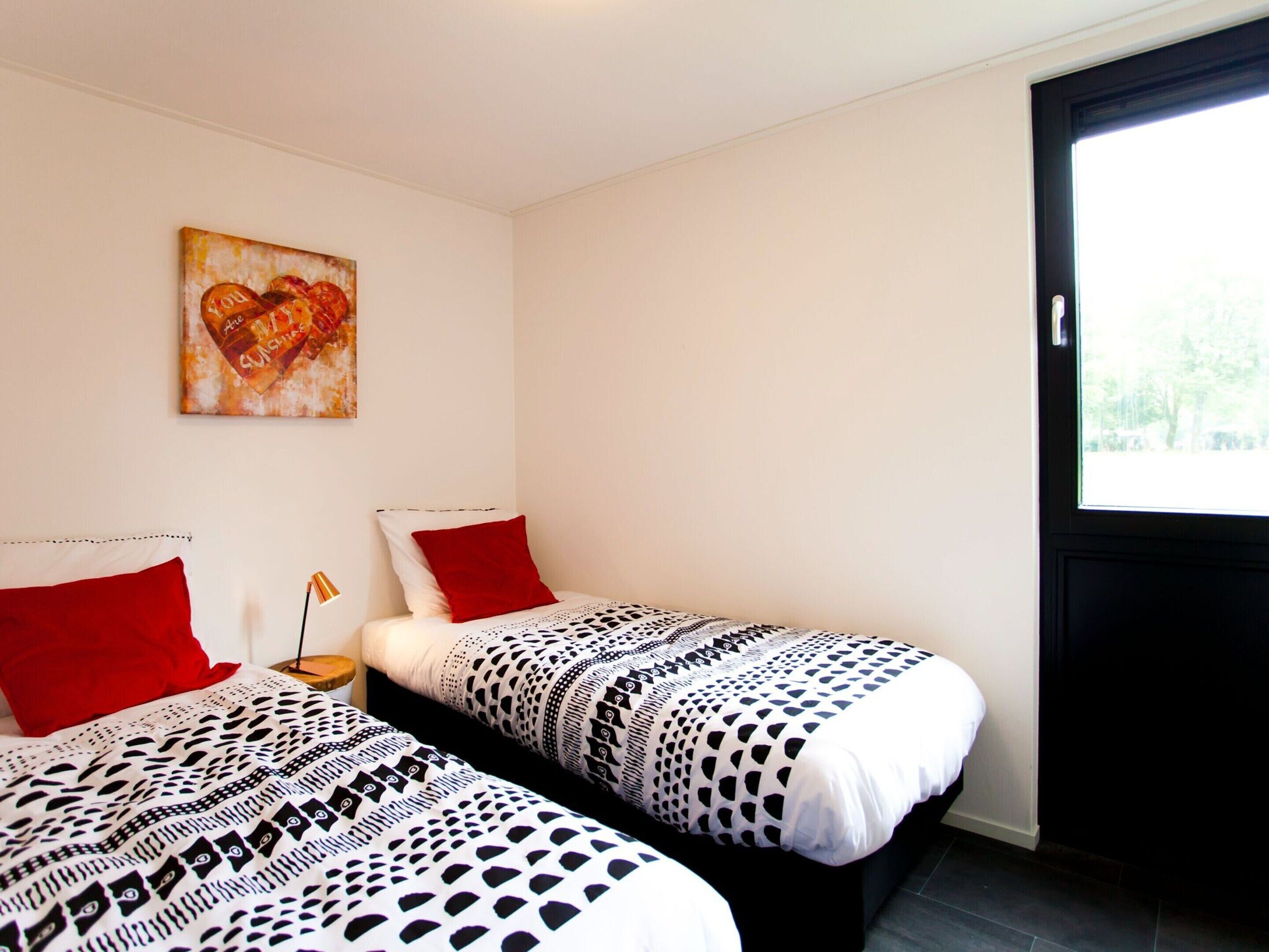 A room with two beds, each with a patterned bedspread and red pillows. There is a painting on the wall and a window with a view of trees.