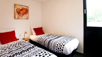 A room with two beds, each with a patterned bedspread and red pillows. There is a painting on the wall and a window with a view of trees.