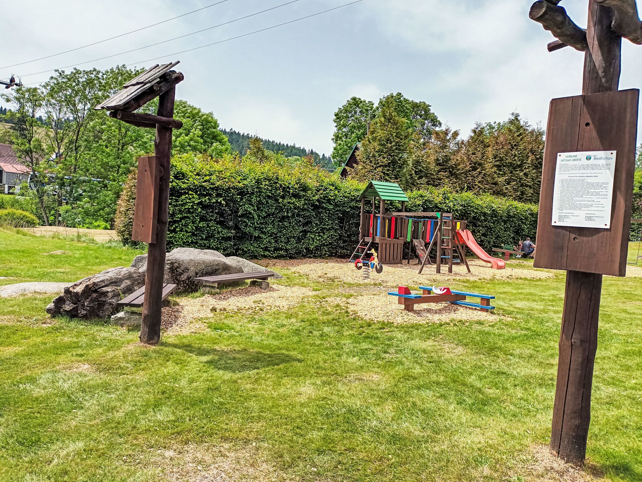 A playground with a slide and climbing structures, surrounded by a grassy area and a wooden fence.