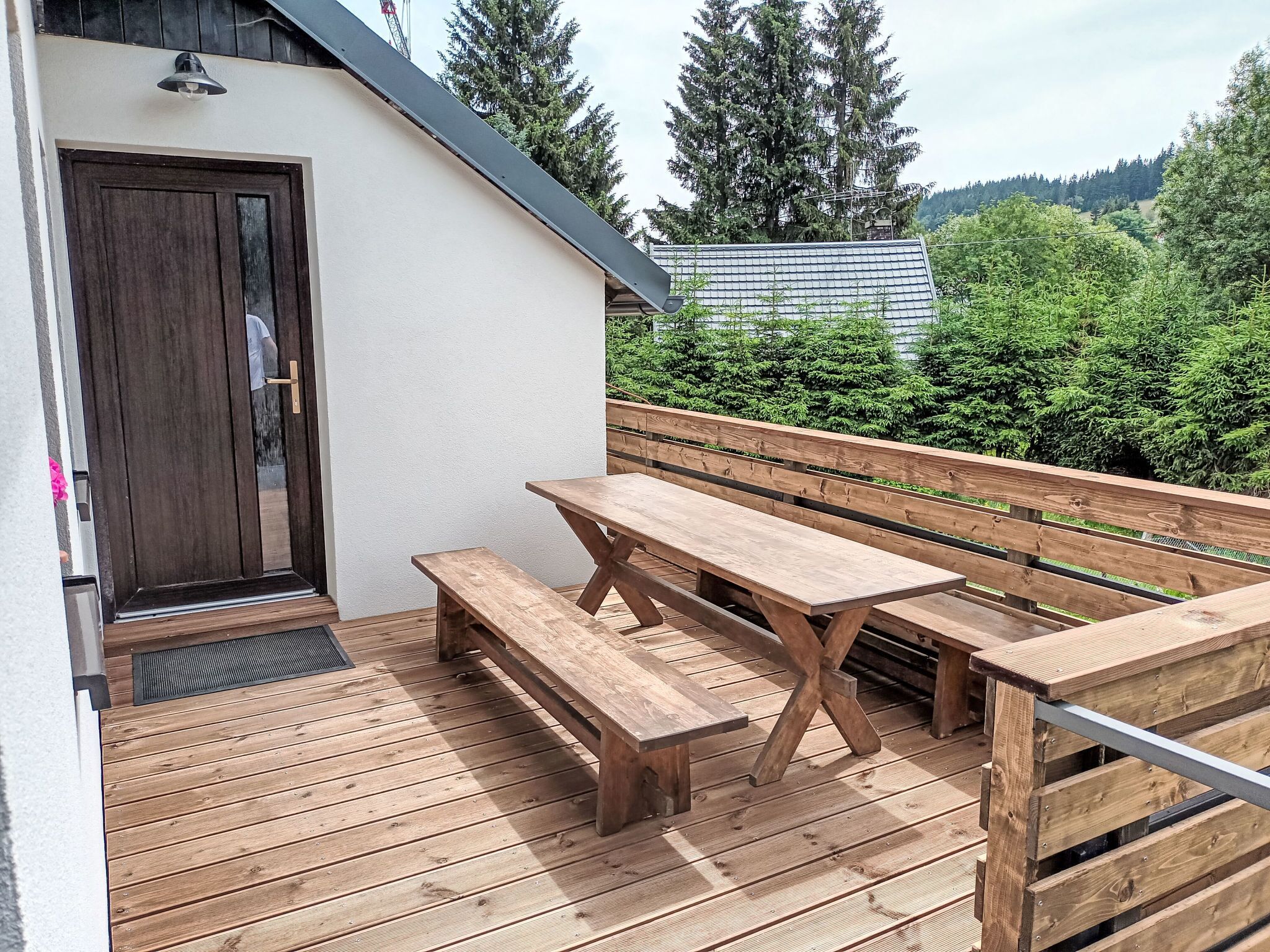 Una terraza de madera con una mesa de picnic y bancos, una puerta, y vistas a árboles y colinas.