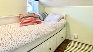 A single bed with a floral-patterned bedspread and multiple pillows, a white dresser with drawers, a window with a view, and a striped rug on the floor. - 5 Star Holiday Home in Borgholm (Borgholm)