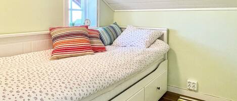 A single bed with a floral-patterned bedspread and multiple pillows, a white dresser with drawers, a window with a view, and a striped rug on the floor.