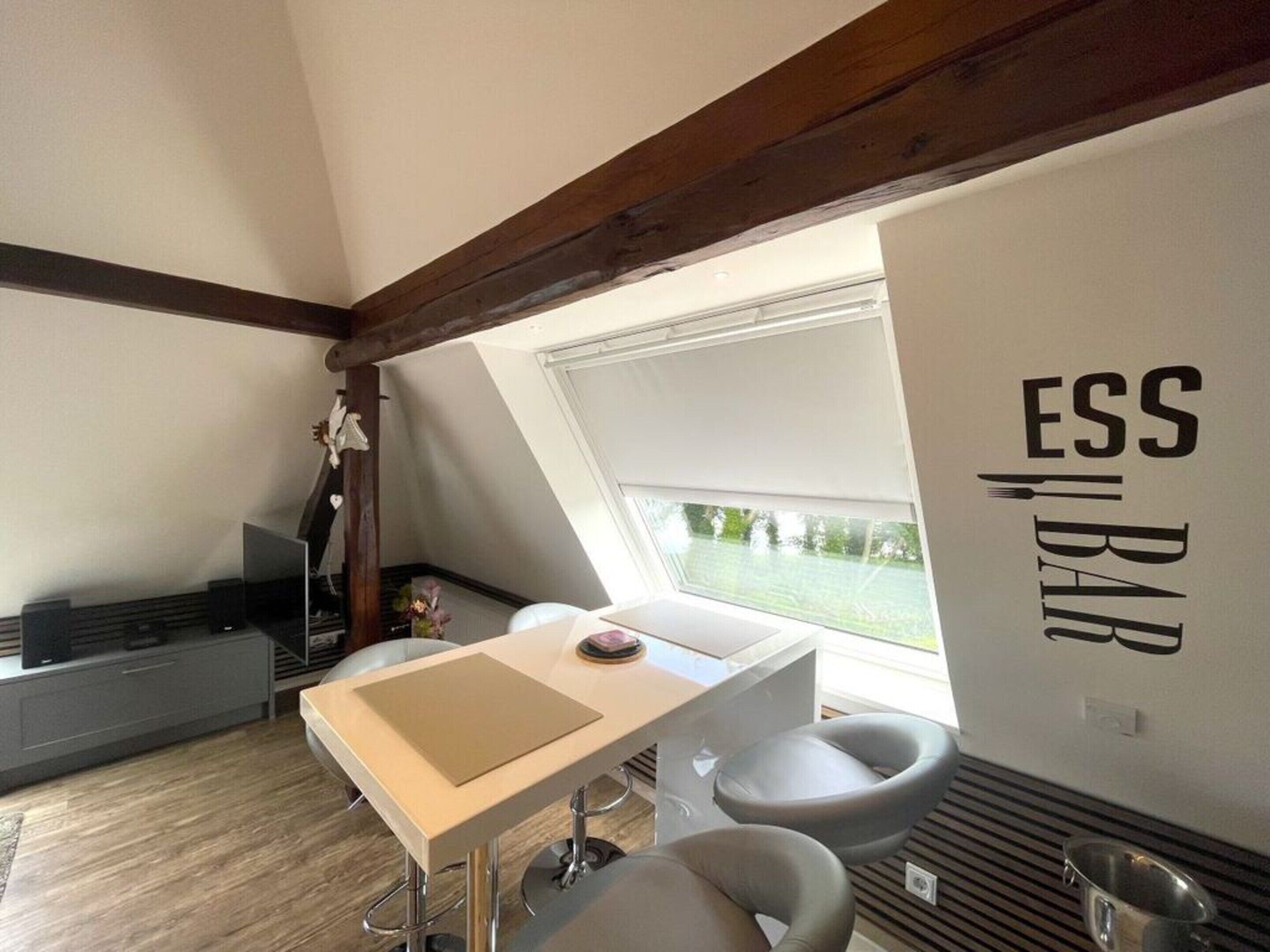 A modern kitchen with a dining area, featuring a white wall with 'ESS BAR' signage, a wooden ceiling, and a view through a window.