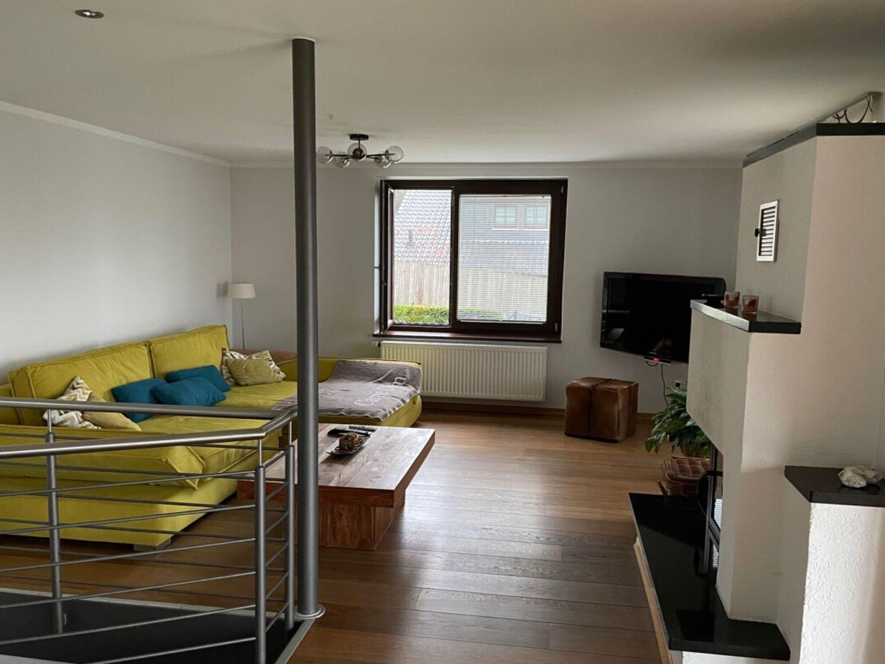 Ein Wohnzimmer mit einem gelben Sofa, einem Holztisch, einem Fernseher auf einem Schrank und einem Fenster mit Jalousien.