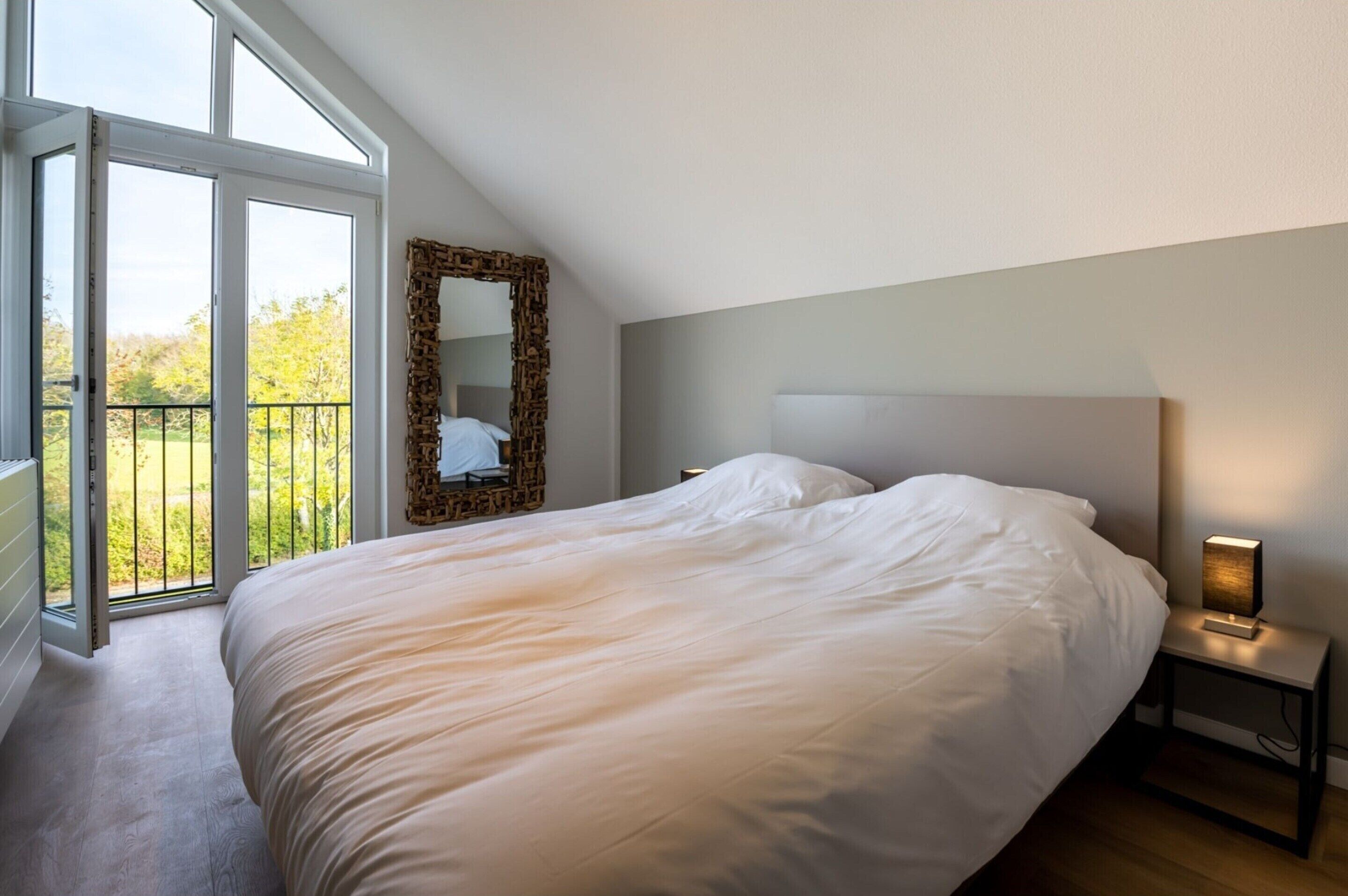 A bedroom with a large bed, a nightstand with a lamp, a mirror, and a balcony with a railing.