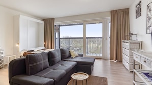 A modern living room with a dark grey sectional sofa, a small round side table, and a view of the outside through large sliding glass doors. - Spacious Studio With Storage Room (Blankenberge)