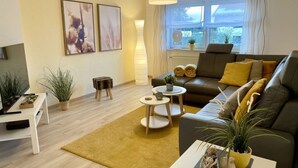 Una sala moderna con sofá, una mesa de centro y plantas en macetas. Se ve una ventana con cortinas y una lámpara.