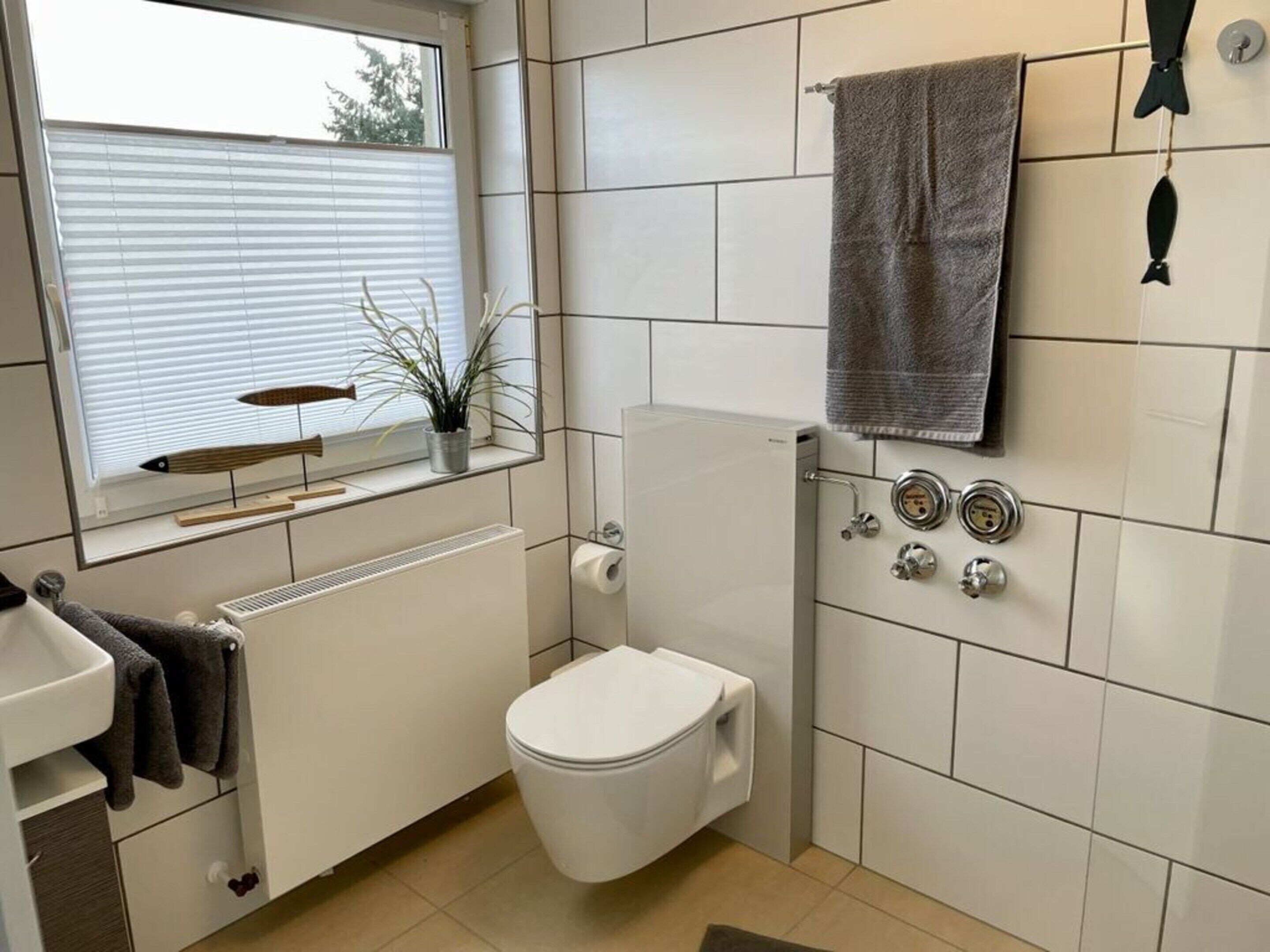A compact bathroom with a toilet, a radiator, a window with blinds, a towel rack, and a plant.