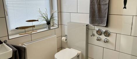 A compact bathroom with a toilet, a radiator, a window with blinds, a towel rack, and a plant.