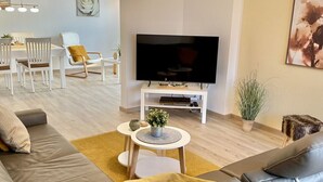 Una sala moderna con un sofá de cuero gris, un mueble blanco para el entretenimiento con un televisor de pantalla plana, una pequeña mesa redonda de centro y un área de comedor al fondo.
