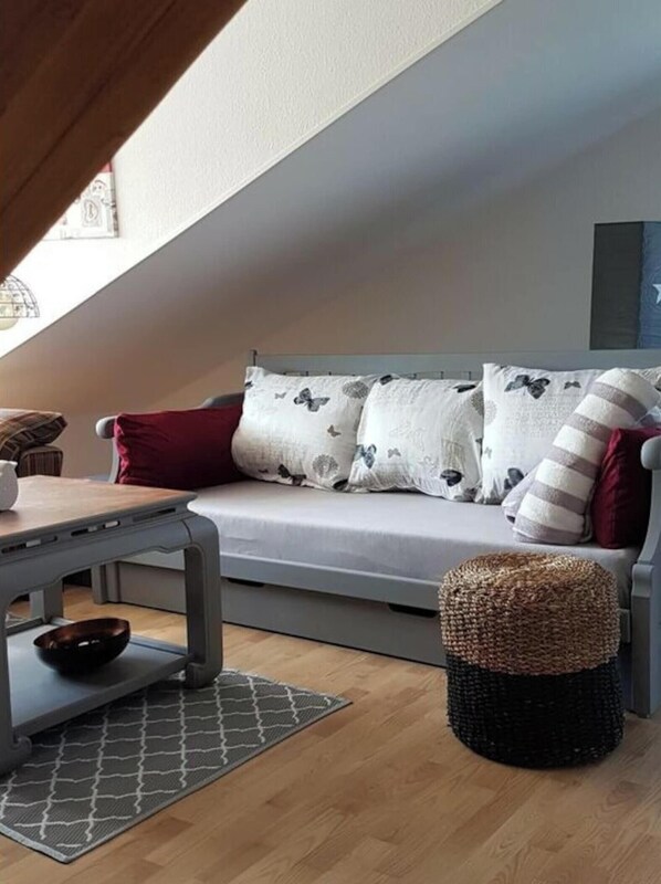 A cozy loft bedroom with a sloped ceiling, a grey sofa with patterned cushions, a small wooden table, and a woven stool. - Apartment in Rieseby (Rieseby)