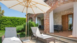 Un patio con sillones de mimbre y una sombrilla grande, un edificio de ladrillo con fachada de piedra y un seto al fondo.