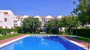 A rectangular swimming pool with a tiled deck, surrounded by greenery and white buildings with red-tiled roofs.