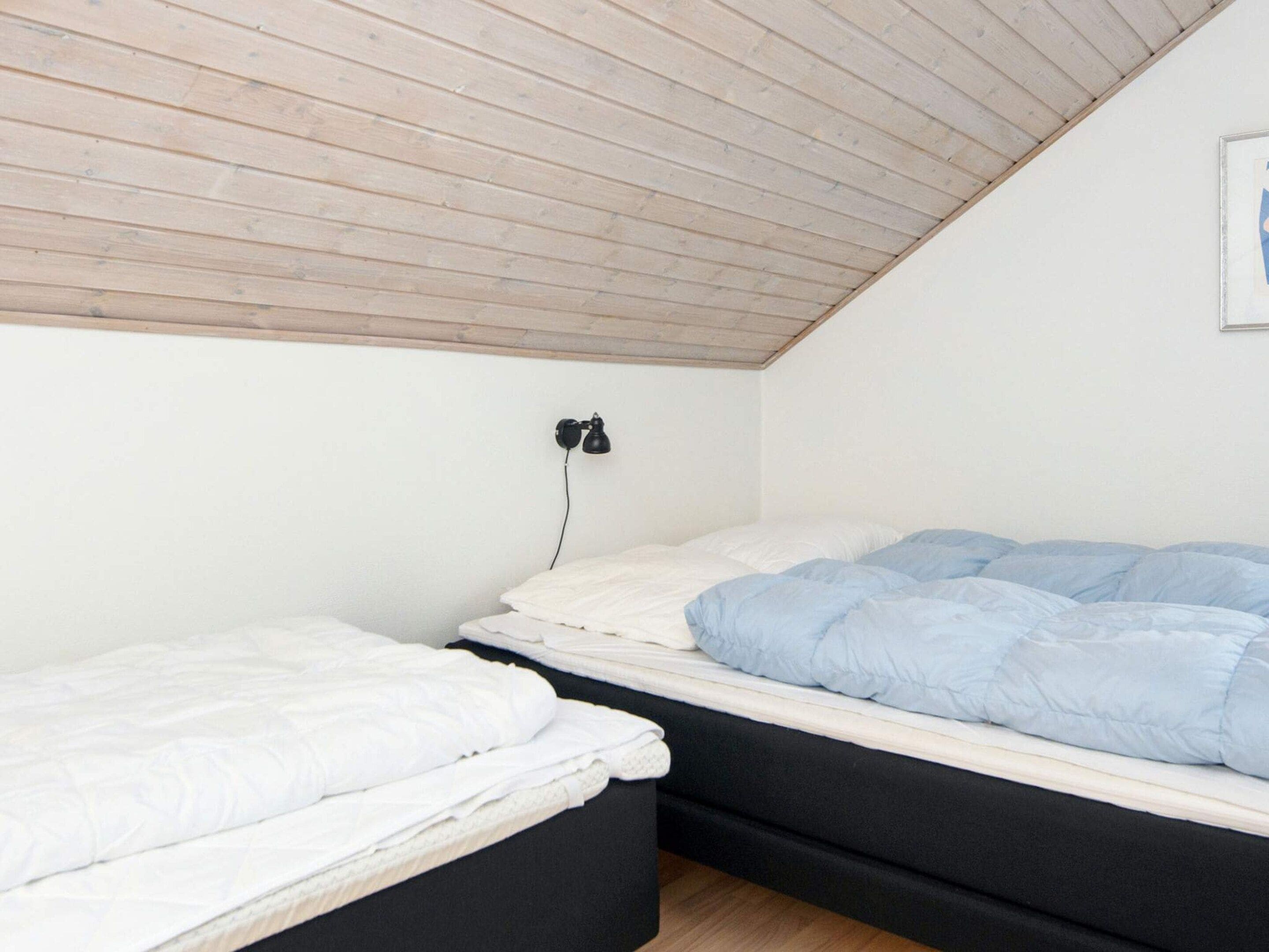 Ein Zimmer mit zwei Betten, eines mit einer weißen und das andere mit einer blauen Matratze. An der Wand ist eine schwarze Wandleuchte befestigt.