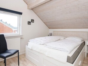 A bedroom with a sloped ceiling, a bed with white bedding, a chair, and a window with blinds. - 4 Star Holiday Home in Romo (Rømø)