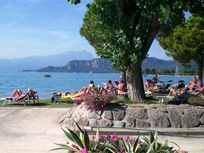 A beachside area with people sunbathing, a large tree, and a stone wall. - Shimmering Serenity in Pastrengo (Pastrengo)