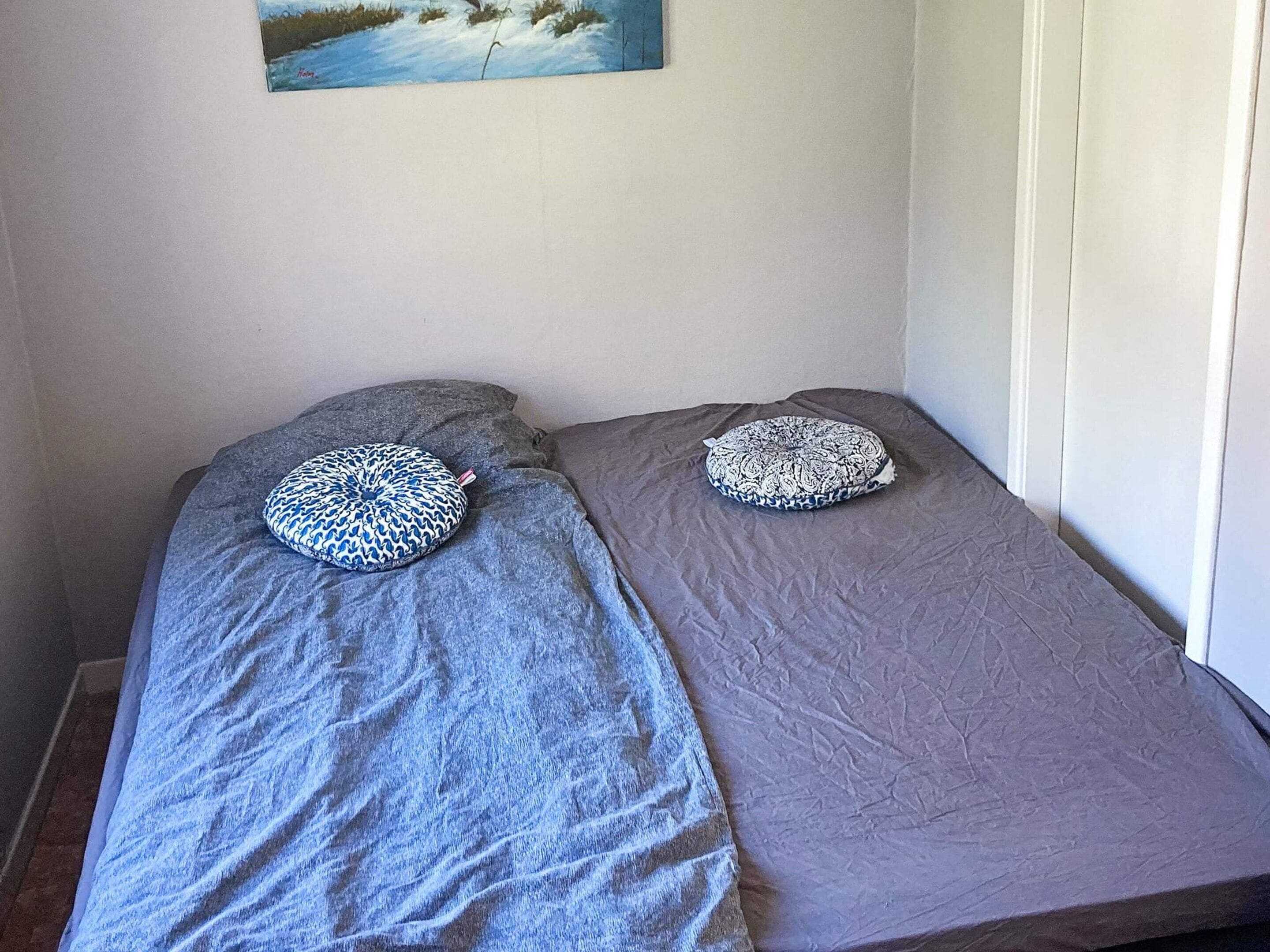 A small room with two single beds, each with a patterned pillow, grey bedding, and a painting on the wall.