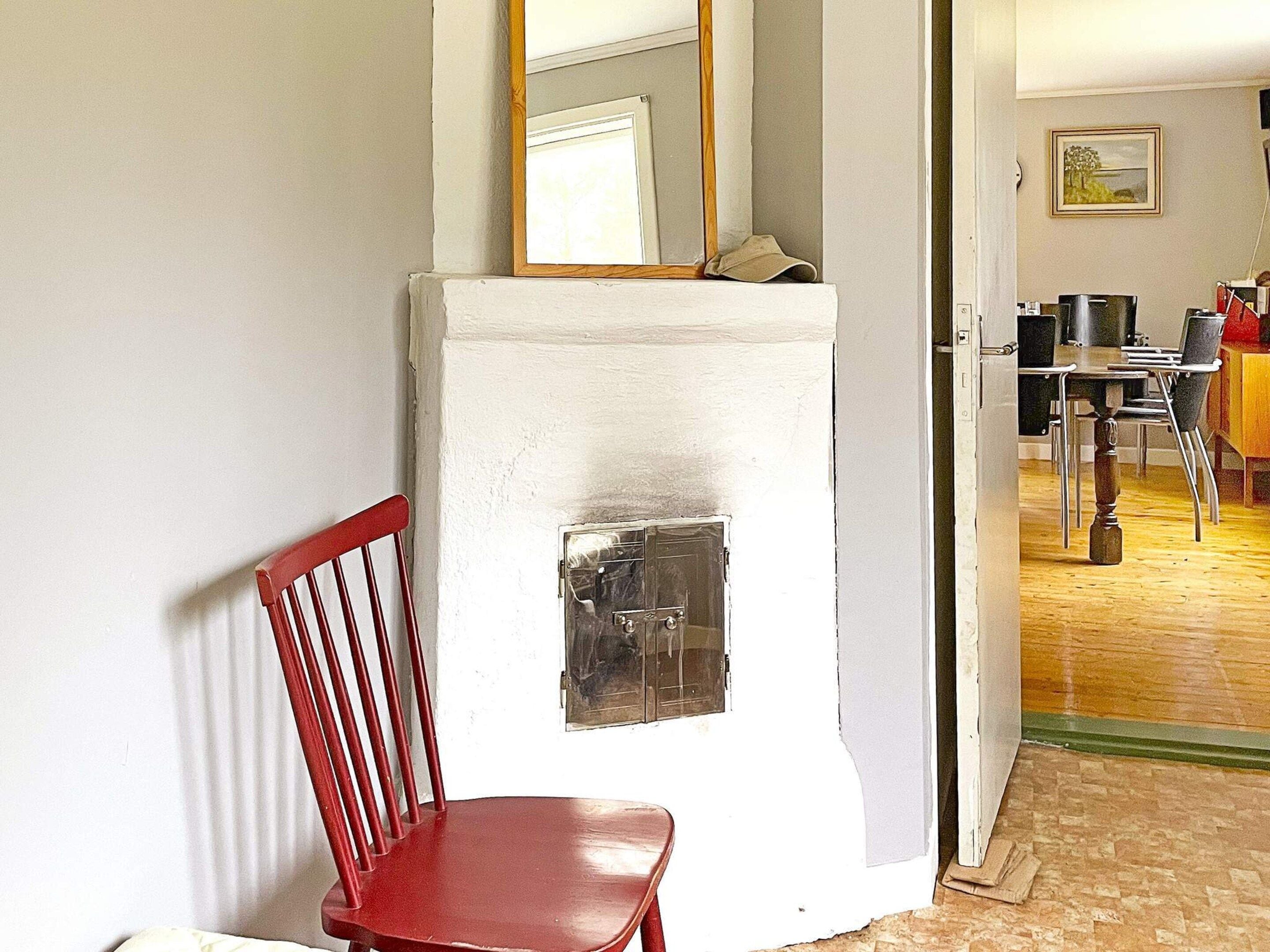 Una habitación con pared blanca, una silla roja, un espejo con marco de madera y una zona de comedor parcialmente visible con sillas y una mesa.