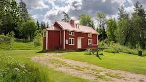 Une maison en bois rouge, avec un toit en pente, entourée d’un champ herbeux et d’arbres.