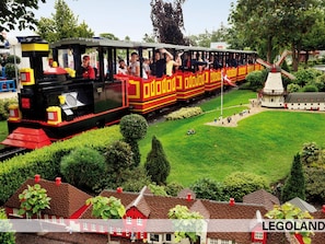 Un train miniature avec des passagers, installé dans un parc où l’on trouve également des maisons miniatures et un moulin à vent.