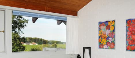 Ein Schlafzimmer mit großem Fenster, einem Bett mit gemustertem Überwurf, einem Nachttisch mit Lampe und zwei Blumenbildern an der Wand.