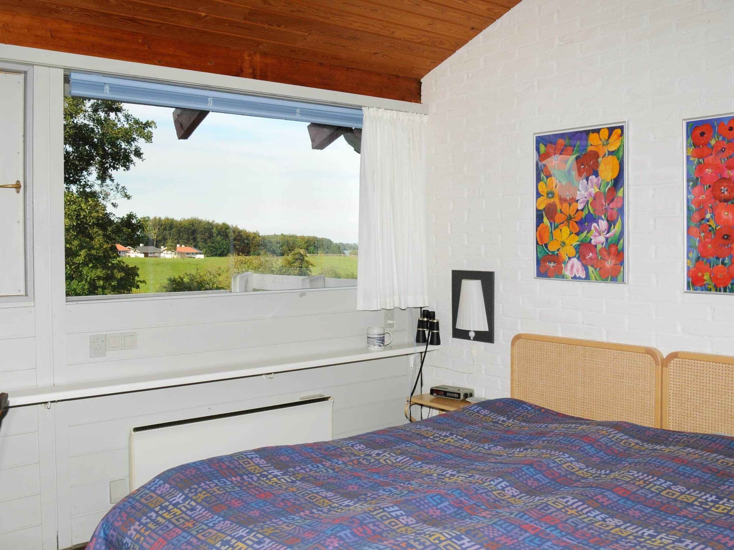 A bedroom with a large window, a bed with a patterned bedspread, a bedside table with a lamp, and two floral paintings on the wall.