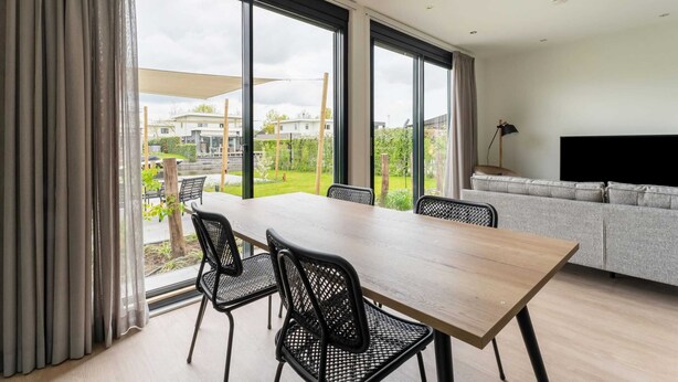 A modern living room with a large window, a dining table with chairs, a grey sofa, and a flat-screen TV. - Amazing Villa in Zeewolde With Terrace (Zeewolde)