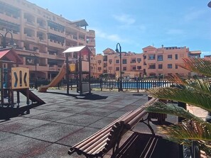 En legeplads med rutsjebaner og klatrestrukturer, omgivet af høje bygninger med altaner.