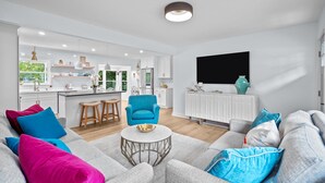 En moderne stue med en grå sofa, farverige puder, et rundt sofabord og en blå lænestol. Et køkken med hvide skabe og et sort tv er synligt i baggrunden.