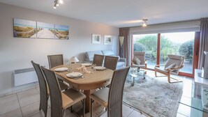 Un comedor con una mesa redonda dispuesta para cuatro personas, rodeada de sillas de mimbre. Al fondo, hay un sofá y una silla, y un ventanal con vistas a zonas verdes.