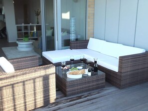 Un espace extérieur moderne, aménagé avec des meubles en osier, une table à plateau en verre et des verres à vin, ainsi qu’un canapé blanc.
