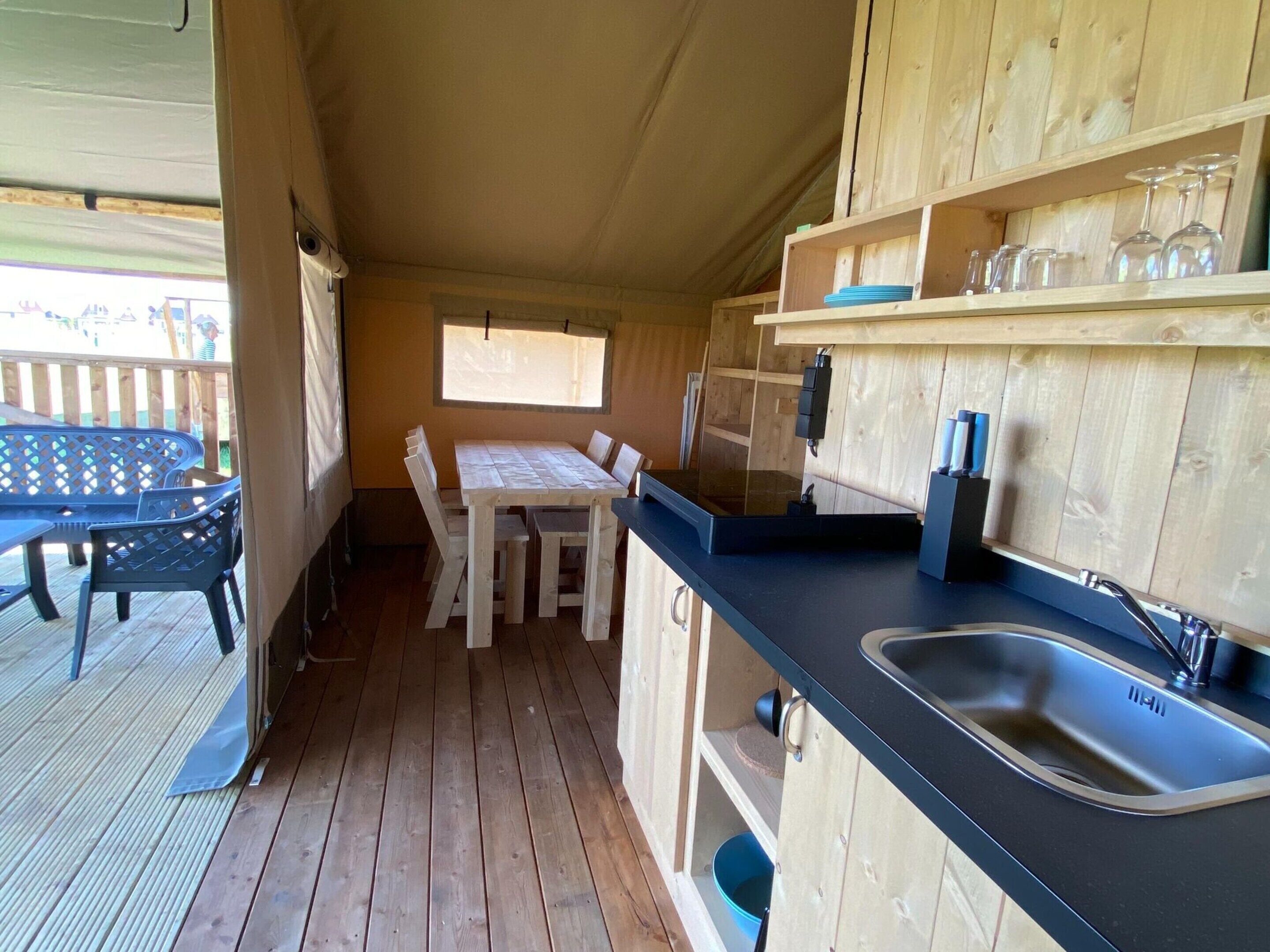 Eine Holzhütte mit Esstisch und Stühlen, einer Küchenzeile inklusive Spüle und Schränken sowie einer Terrasse mit Tisch und Stühlen.