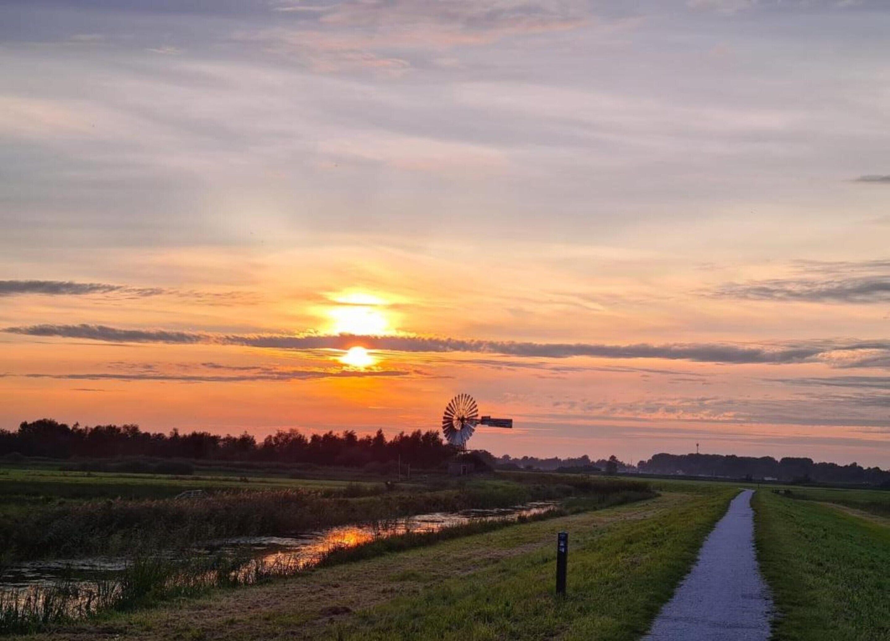 A sunset over a rural landscape with a windmill and a narrow water channel.