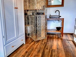 Ein Zimmer mit einem weißen Kleiderschrank, einem dekorativen Metallteiler, einem Holztischchen mit Waschbecken und Holzboden.