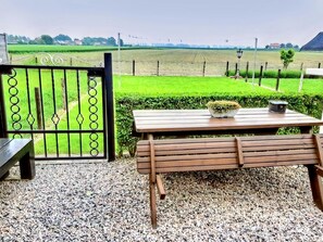 Eine Terrasse mit einem Holztisch und einer Bank, einem Blumentopf und Blick auf ein grünes Feld durch ein Metallgitter.